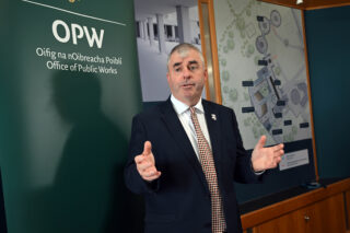 a man stands with his hands spread out, with an OPW logo on the wall behind him