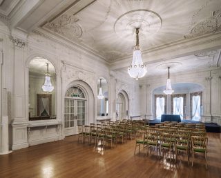 The Ballroom is lit by multiple chandeliers hanging from the ceiling. The wooden floor shines with the light from above. Chairs fill the front space of the room facing the stage, framed by a tri-window