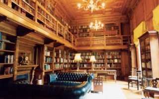 The library is bathed in golden light from the candelabras on the ceiling. Books line the room over two floors, the second floor is a balcony level overlooking the lower which is furnished with two black leather sofas.