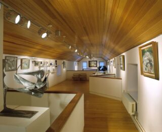 Interior image of gallery with paintings hung on white walls and a metallic sculpture in the foreground.