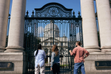 3 people stand outside closed black gates looking at the exterior of the government buildings