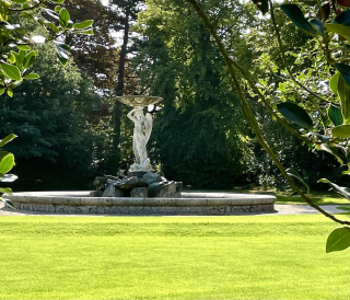 A female statue stands in the centre of a fountain with trees framing the edges & background of the image. In the foreground, the grass is lush and bright green in the bright day.