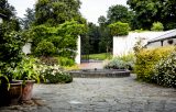 The courtyard is framed by various plants and bushes in different shades of green, with a circular fountain in the centre and open gates to the background leading into the main park area