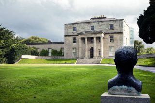 In the right foreground of the image the back of the bust of Patrick Pearse can be seen looking at the front exterior of the Pearse Museum. The grey clouds dominate the background of the image.