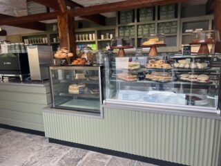 A café display of cakes and sandwiches alongside a modern coffee machine. The counter is painted sage green. There are exposed wood beams above. The space is cosy. The floor is raw flagstones. A cosy and rustic style.