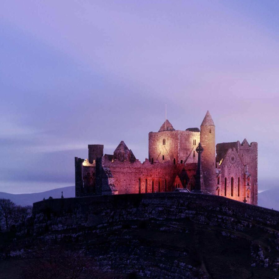 Rock of Cashel lit up at night. The sky appears purply blue and the rock beneath the Castle is cast in shadows.