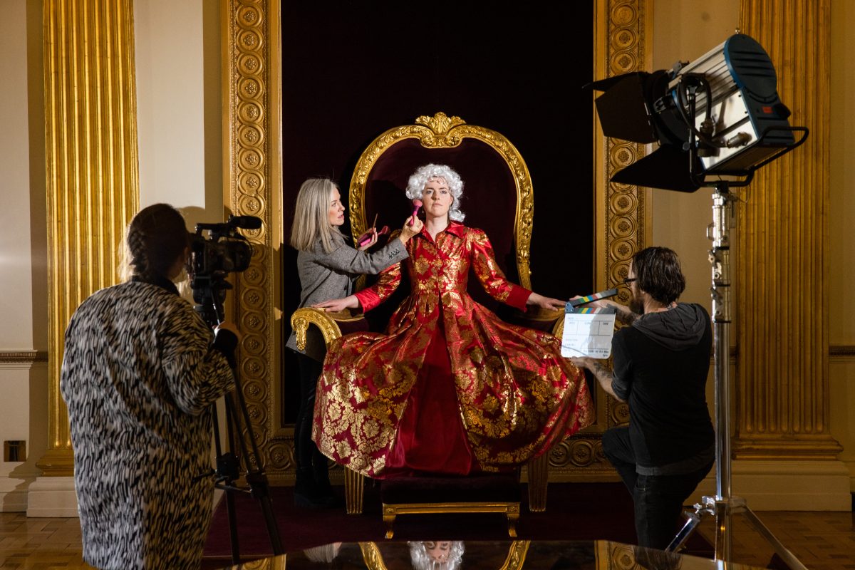 Photo of a woman in costume on the throne having make-up applied as a cameraman and production assistant waits to resume filming work.