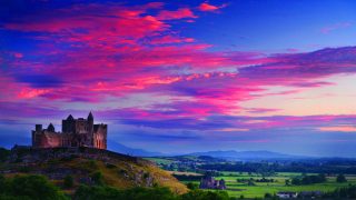 The Rock of Cashel stands on the left dimly lit by a spectacular sunset sky with purple and pink clouds