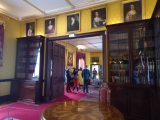 Within Kilkenny Castle we see a group turned towards a guide. They are visible through an open doorway. Above the door there are portraits. The walls are covered in a sunshine-yellow wallpaper with an embossed pattern. The carpet carpet and reds are a rich wine-red colour. There are glass cabinets full of old books and a round wooden table highly decorative and varnished.