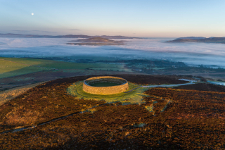 A ring fort is lit by the sunrise. It is surrounded by marshy plant-life coloured in browns and greens. The sky in the background is a yellowy-blue and is misted in the early morning