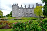 The exterior of Kilkenny Castle, the photo has been taken in the rose garden, grass and pink roses sprawl on the ground in front of the Castle.