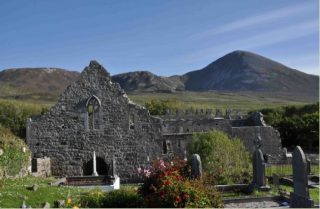 The remainders of Murrisk Abbey lie surrounded by graves and in the shadow of Croagh Patrick, which looms large in the background. It is a bright day, with no clouds in the blue sky.