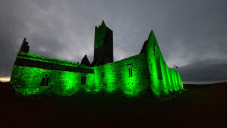 The Rosserk ruins are floodlit in green for st. patrick's day. The clouds are a slate grey in the background, and the landscape to the front of the ruins is completely black in the dark