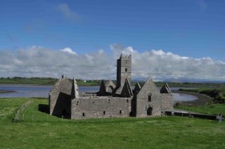 Ruins of a medieval monastery lie on the banks of the River Moy in Co. Mayo. The grass surrounding the ruins is lush and green, while the sky behind it is blue, despite the clouds