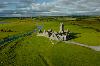 A stone abbey sits in the middle of a green field with a river flowing past on its left side