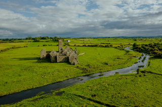 A stone abbey sits in the middle of a green field with a river flowing past on its right side