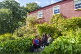 Three young people sitting in front of Glebe House and Gallery. They are surrounded by plants and foliage. Glebe House stands behind them