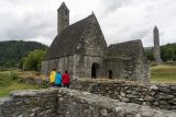 St. Kevin’s Church better known as St. Kevin’s Kitchen is a nave-and-chancel church of the 12th century. There are people in yellow, blue and red jackets outside the church. There is a stone wall in the foreground