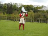 An image of a living history display actor. He is dressed as a solider, wearing a red and yellow uniform. He is shooting a gun.