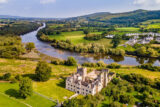 aerial view of the manor house, with its tower ruins behind it, and a view of the surrounding hills and river at the back.