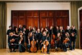 A group of musicians from the Irish Baroque Orchestra stand in a group in front of large mahogany doors. The musicians hold their instruments- two cellists it at the front, holding cellos with their thighs. Violinists, trumpeters, violists and one double bassist stand and sit around the cellists. Members of the group look jovially at each other and the conductor sits in the middle, smiling at the camera.