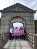 6 guides pose in front of an arched gate holding a flag that promotes National Heritage Week