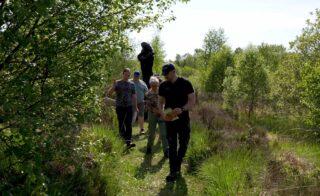 A group of people participates in a guided walk at Corlea Trackway during Biodiversity Week. The guide, holding a book, leads the group along a lush, green path surrounded by trees and tall grass.