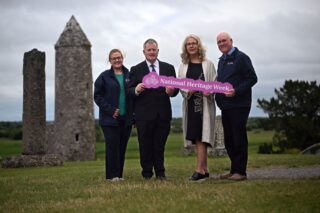 Four people stand holding a pink and white sign saying National Heritage Week. They are outdoors in at Clonmacnoise Monastic Site. Behind them are the round tower and other remains of the monastery. They stand on green grass with a cloudy sky behind.