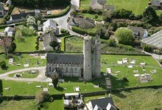 An aerial view of a round tower and church surrounded by green grass and local roads.