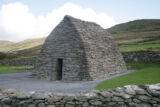 A front-facing view of Gallarus Oratory, a stone building shaped like an upturned boat, backdropped by the mountains