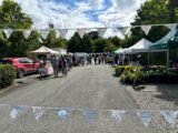 Oldbridge Farmers Market, bunting decorates the street and stretches across the image. Colourful tents line the street as people peruse and shop.