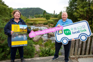 A woman and a man stand holding signs advertising heritage week and the local bus links to Glendalough. They are standing in front of the backdrop of the mountains and monastic site.