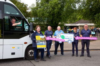 5 people are standing holding signs advertising heritage week and the local links bus to Glendalough. They are standing in front of the visitor centre. There is a bus parked to their left with a driver giving a thumbs up out the window