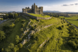 A castle perched on top of a craggy green hill, with the mountains and blue sky as its backdrop