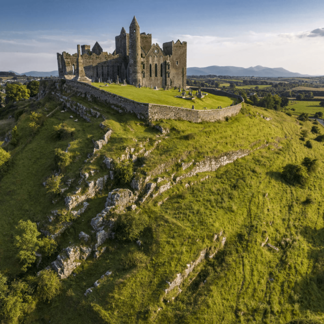 A castle perched on top of a craggy green hill, with the mountains and blue sky as its backdrop