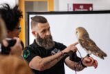 John Carrig, a man with a beard and hair tied up in a bun, holding a barn owl. The owl is sitting calmly on his hand while John looks at her and talks. There are some people on the left side of the photo watching the owl curiously.