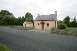 View of the cottage from a side angle, and there is a small 'shed-like' building next to it, as they stand by the side of the road