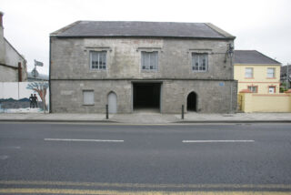 front elevation of a grey brick house, with a yellow painted building to its right, and a wall mural on the left.