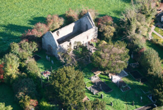 aerial view of the roofless church, situated within a graveyard and trees surrounding the ruins