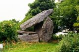 3 stones prop up a larger, horizontally slanted cap stone. Trees grow around the portal tomb.