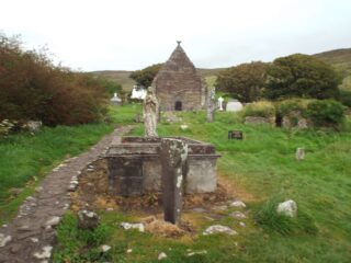 a stone well, with a statue of Mary sits in the foreground, with the church in the back distance