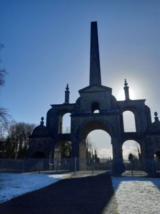 a portrait view of the conolly folly backlit by the low sunlight, with frost covering the grass in front of the monument