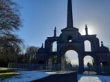 a multiple arched structure with an obelisk pillar in the centre, backlit by the sun