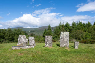 Six standing stones in a field backdropped by Evergreen trees. Five of the stones are standing, and of differing heights, and one is horizontal.