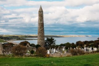 the round tower stands among the graves in the cemetery, with the sea visible just behind it.