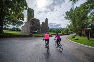 Two women cycle the road leading to the abbey. Backdropped by the cloudy sky, the abbey stands large.