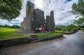 external view of the abbey, the sun straining to come through the clouds behind it. Two women approach the entrance, reading information panels just outside the doorway.