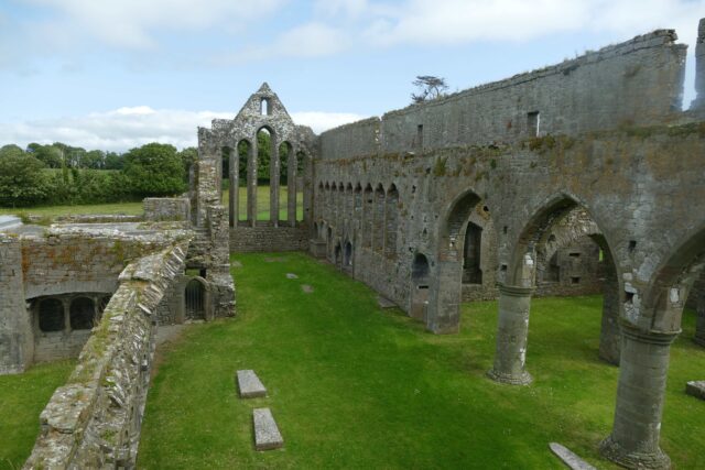 the roofless remains of the friary. Archways of the courtyard are to the right, with the now glassless pointed window is at the end of the courtyard.