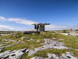 the stone tomb in the distance, the blue sky as the backdrop, and the rugged, rocky landscape of the Burren in the foreground.