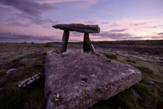 a large horizontal stone lies in the foreground, with the portal tomb standing just behind it, backdropped by the purple evening sky.